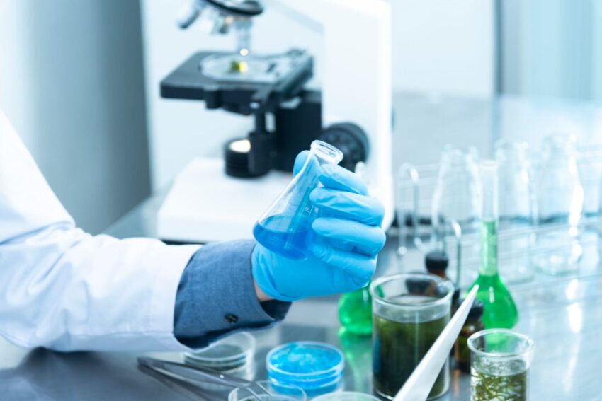 Gloved hand in a lab coat holding a flask of bright blue liquid beside petri dishes and a microscope