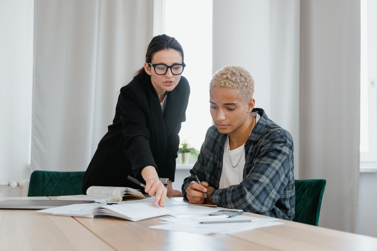 Tutor in a black blazer pointing at open textbooks while a student takes notes at a bright desk